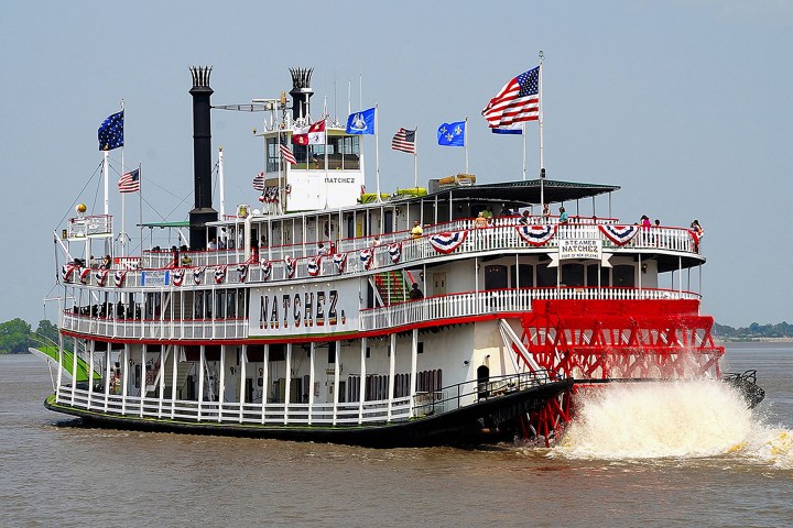 Paddle steamboat with flags on river, named Natchez, churning water with a red paddlewheel.