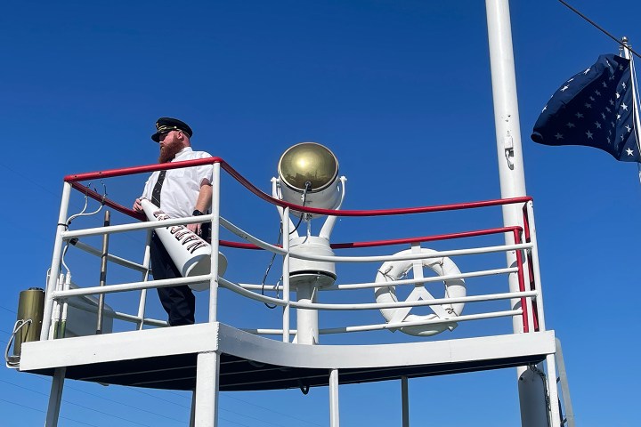 Boat captain stands on deck with megaphone, blue sky background, starry flag in view.