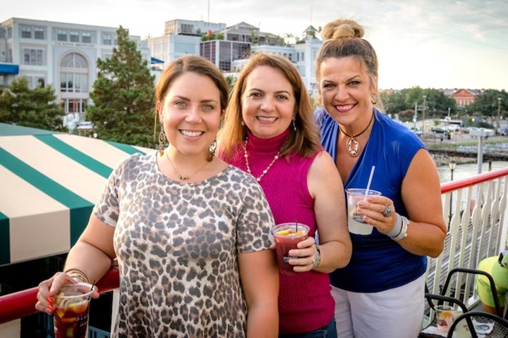 Three women smiling and holding drinks on an outdoor patio with buildings in the background.