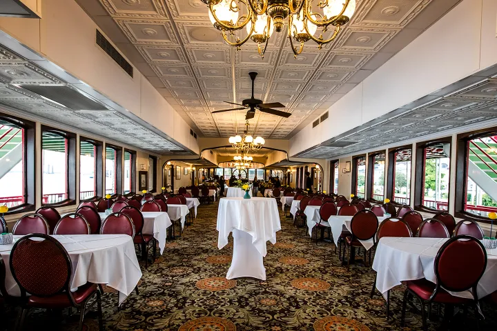 Elegant dining room with chandeliers, white tablecloths, and red chairs on a patterned carpet.