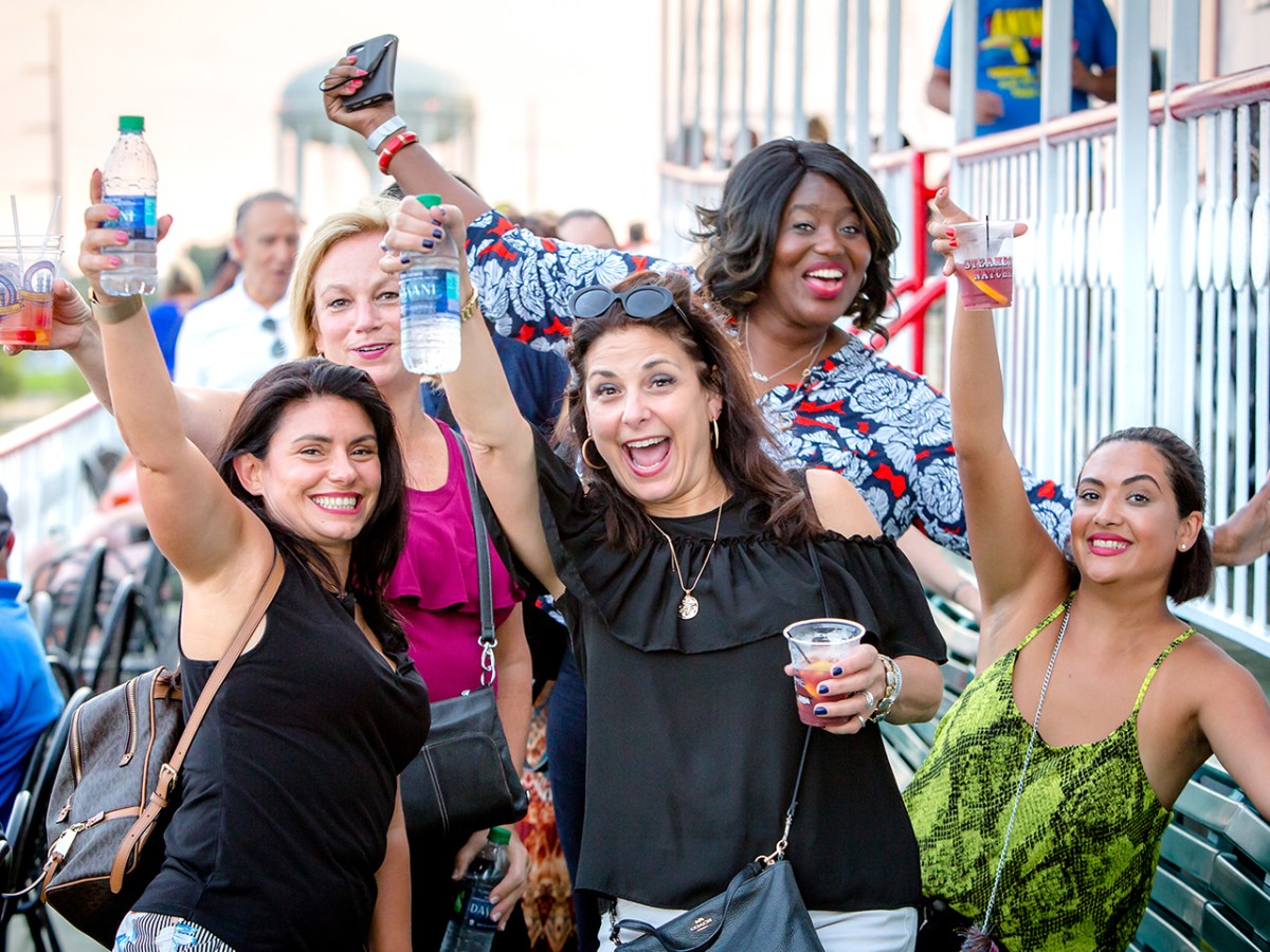 Five women smiling and cheering, holding drinks outdoors.