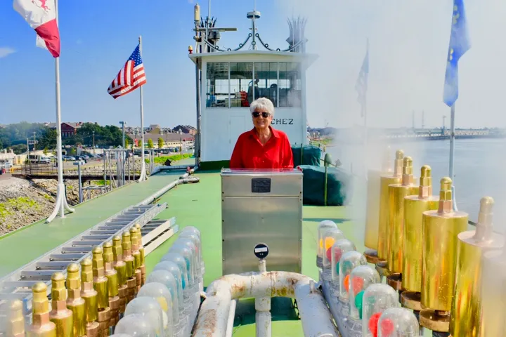 Person in red shirt stands on a ship deck with brass fixtures and flags in the background.