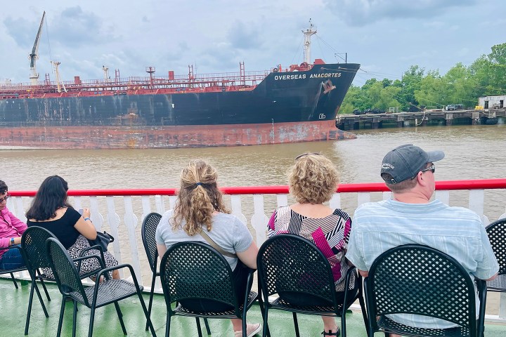 People sitting on a boat watching a large ship named 'Overseas Anacortes' pass by on a river.