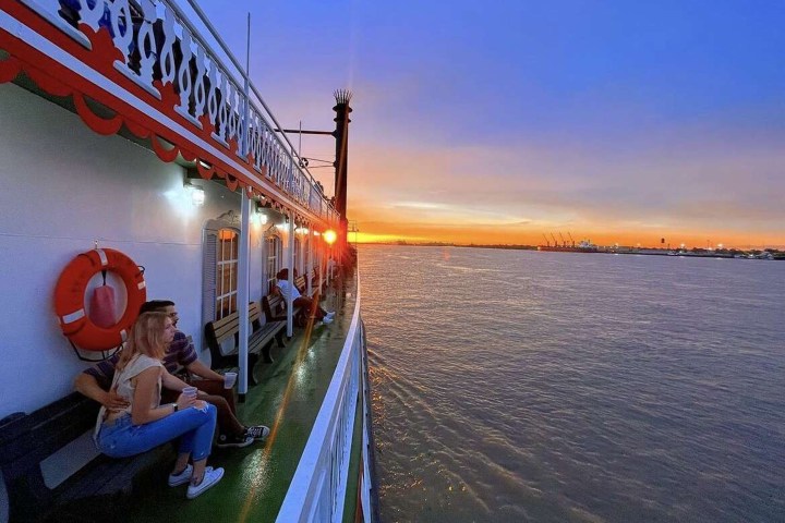People on a boat enjoy a sunset over the water with a calm sky and distant city lights.