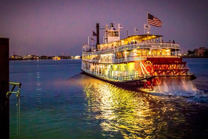 Paddle steamboat on a river at dusk with city lights in the background.