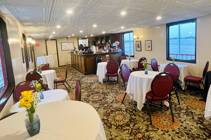 Cozy café with round tables, white tablecloths, and yellow flowers, overlooking water through large windows.