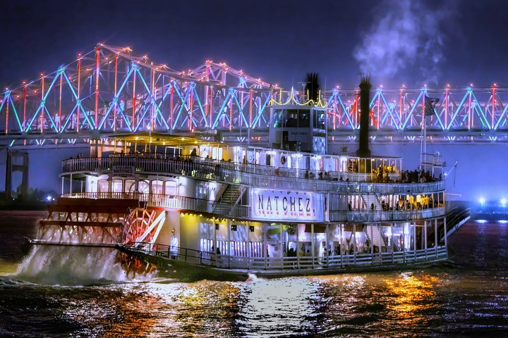 Illuminated riverboat Natchez on water at night with lit bridge in background.