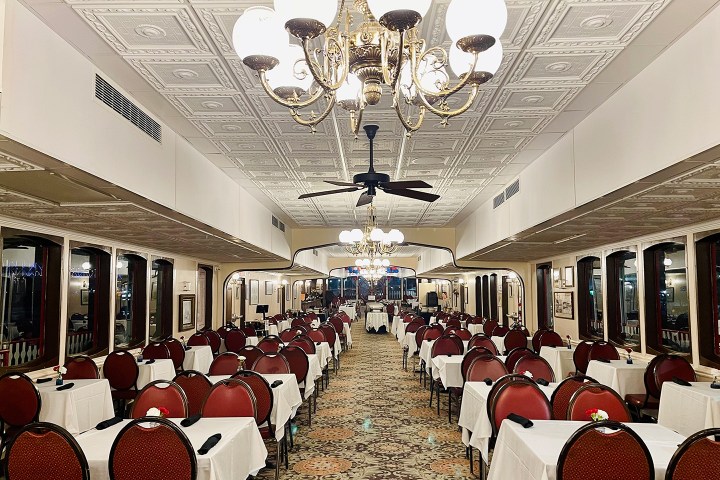 Elegant dining room with red chairs, white tablecloths, chandeliers, and patterned carpet.