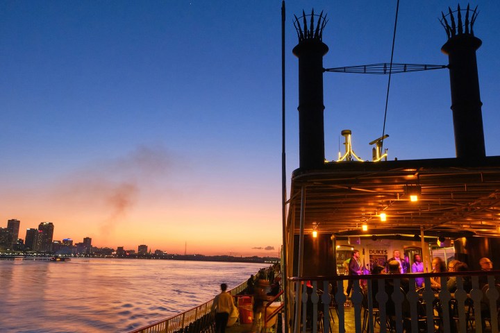 Steamboat on river at sunset with city skyline and orange sky in background.