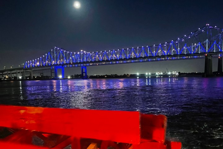 Night view of a bridge lit in blue with full moon above and water reflection below.