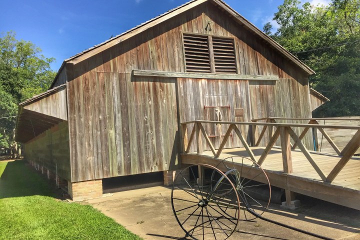 Wooden barn with large doors and wagon wheel, surrounded by greenery on a sunny day.