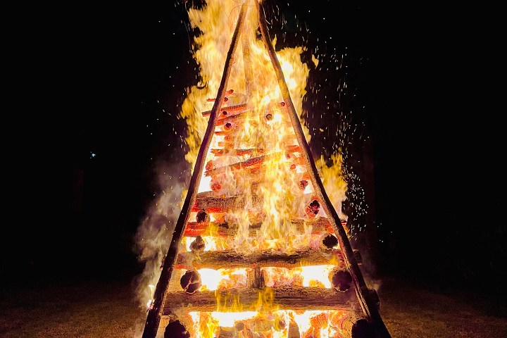Large triangular bonfire with stacked logs burning brightly at night.
