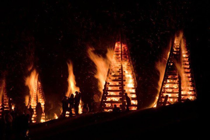 Silhouetted people near large triangular bonfires at night.