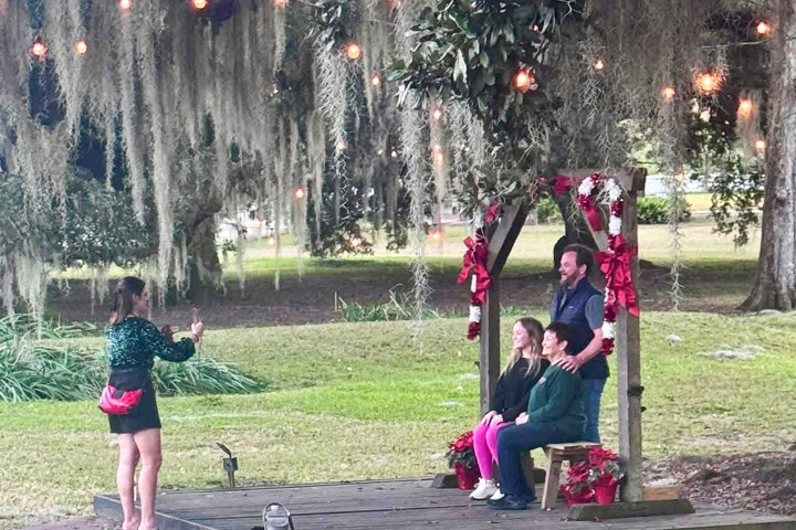 Woman photographs group on decorated outdoor stage with lights and greenery.