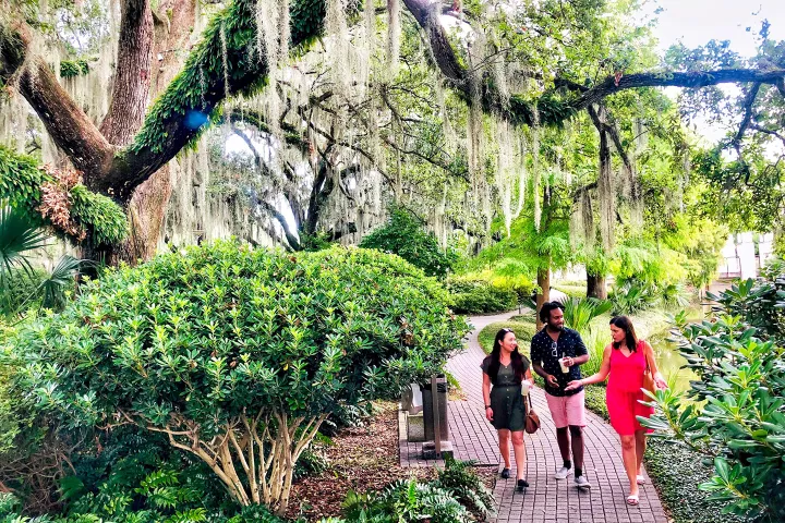 Three people walk on a brick path under mossy trees in a lush green garden.