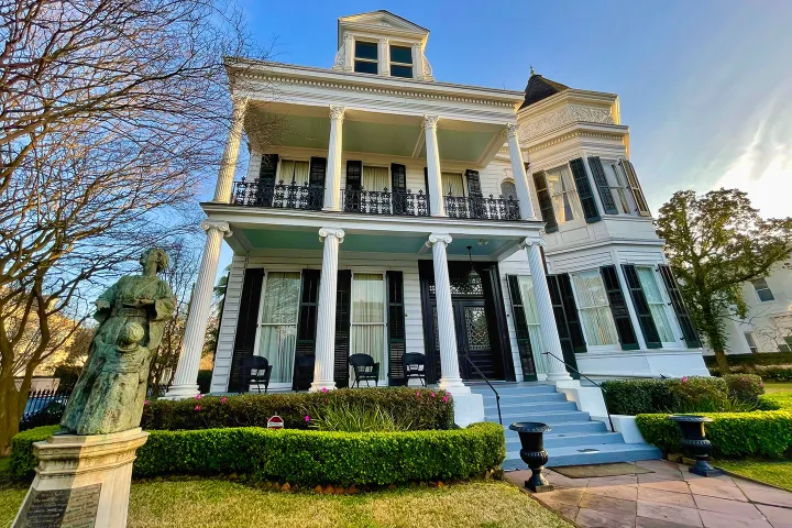 Three-story white house with columns, ornate balcony, and front statue in garden.