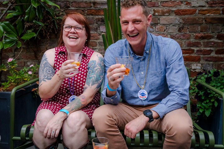 Two people sitting, laughing, holding drinks against a brick wall background.