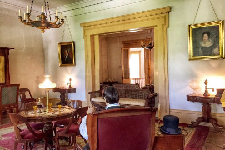 Vintage-style room with chandelier, portraits, furniture, and a person facing away sitting at a table.