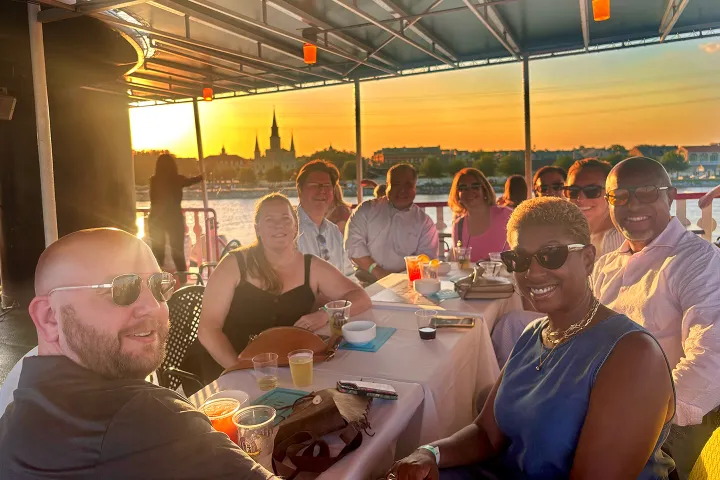 Group of people sitting at a table on a boat, smiling at sunset with a city skyline in the background.