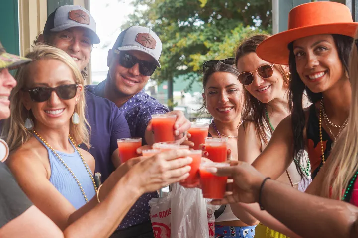 Group of people toasting with red drinks, smiling and wearing sunglasses and hats outdoors.