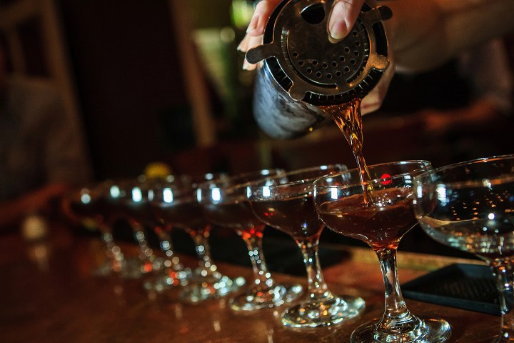 Bartender pouring cocktails into a row of glasses on a bar counter.