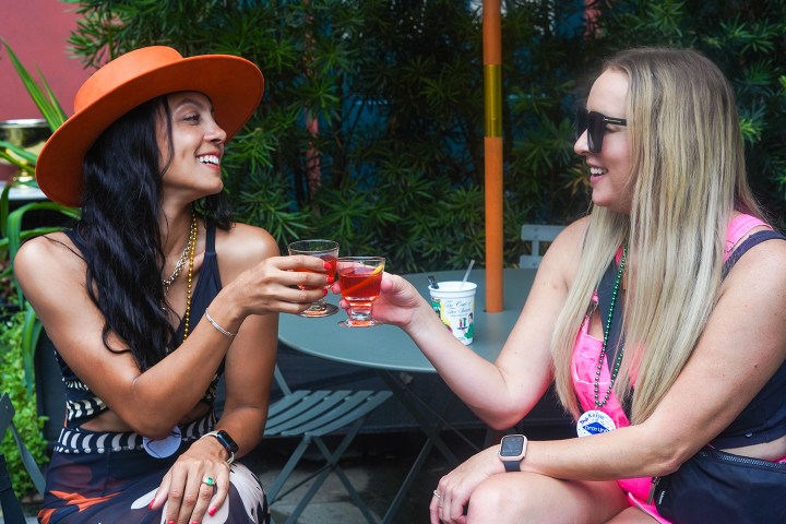 Two women clinking glasses outdoors, one wearing an orange hat, the other with sunglasses.