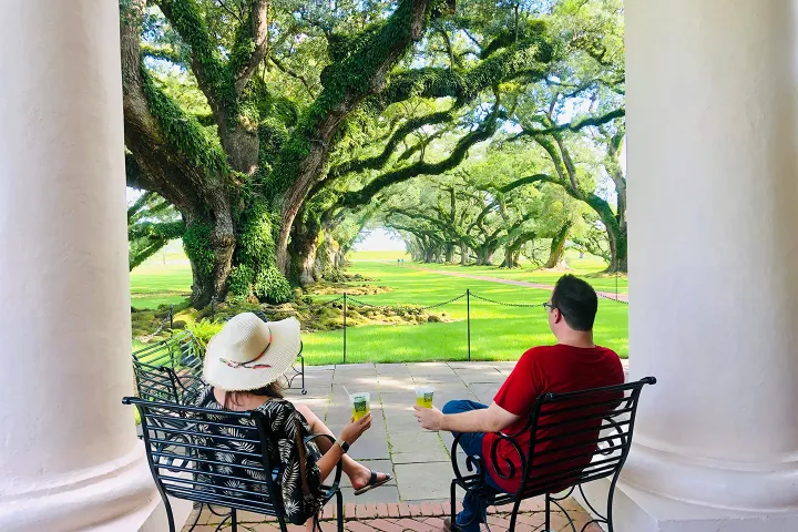 Two people sitting on chairs under large trees, holding drinks, framed by white columns.