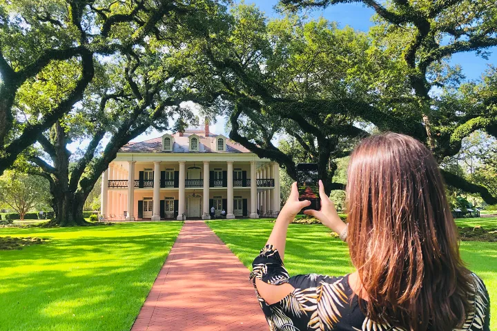 Woman photographing a large, historic house with columns and trees in the background.