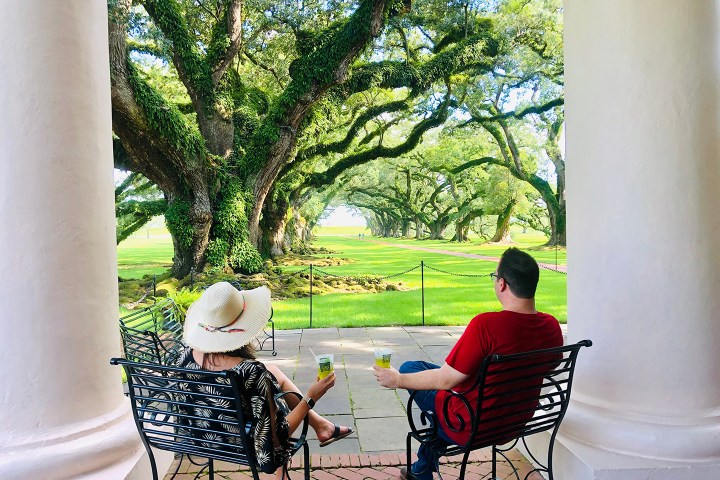 Two people seated on a veranda, facing large oak trees with sprawling branches.