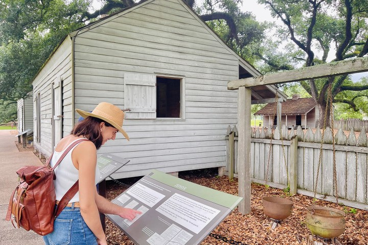 Woman in hat reads sign by wooden cabin and fence with trees in background.