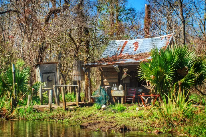 Rustic wooden shack with tin roof, surrounded by trees, near a small riverbank.