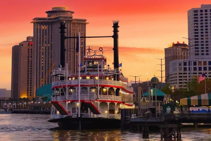 Riverboat docked by city buildings at sunset with vibrant sky.