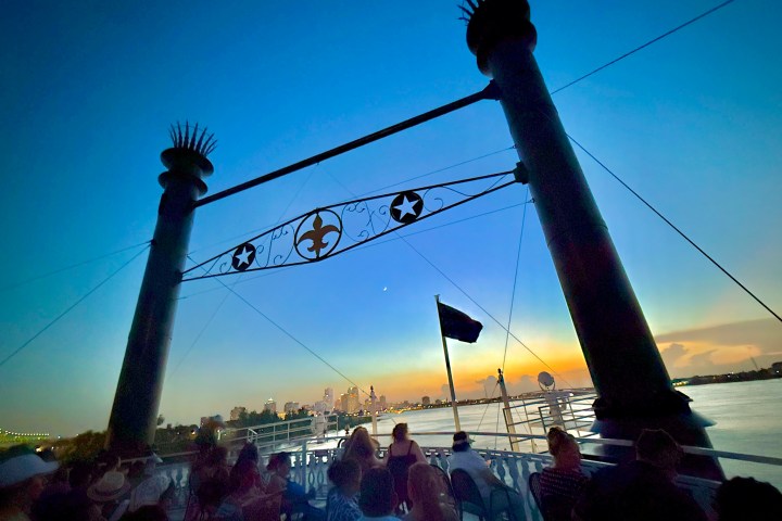 Silhouetted people on a boat deck at sunset with city skyline and decorative arch.