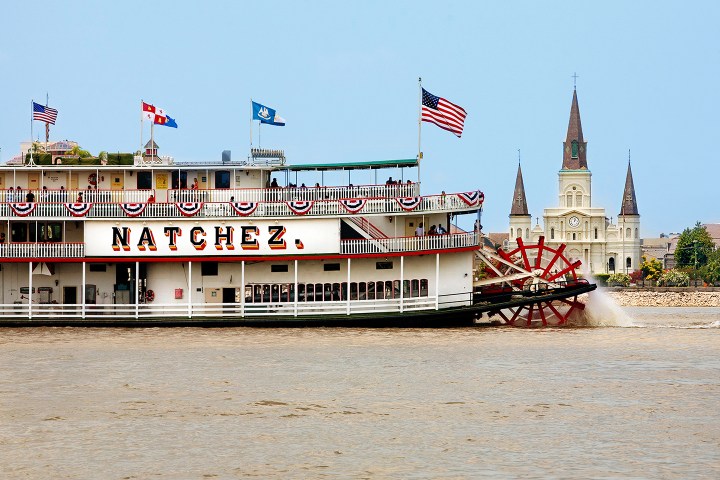 Riverboat 'Natchez' sailing with flags and cathedral in background.