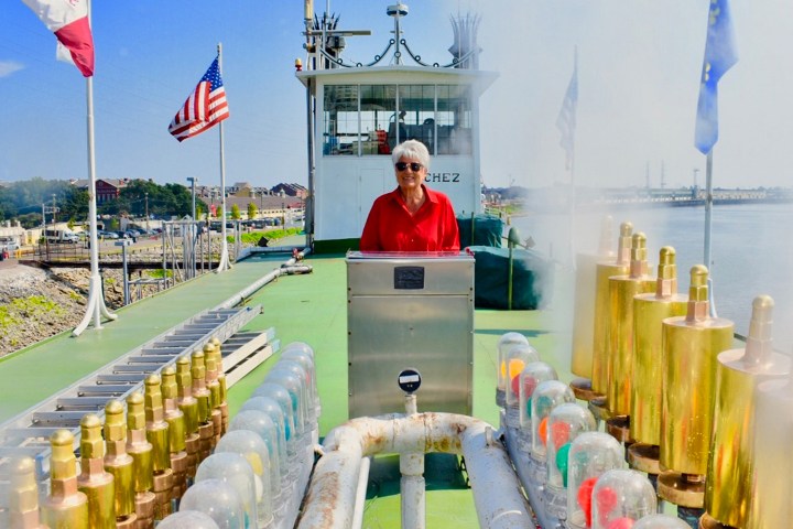 Person in red shirt stands on riverboat deck with flags and smokestack in the background.