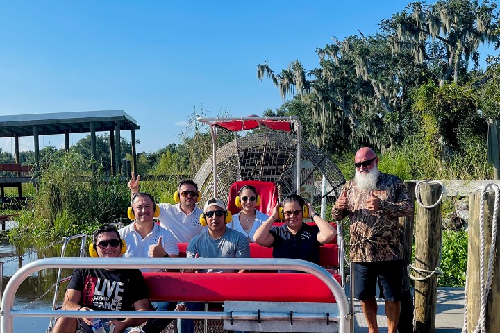 Group of people on an airboat with trees and dock in the background under a clear blue sky.