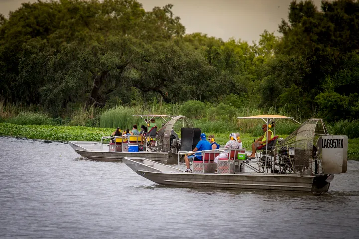 Two airboats with people navigating a calm river beside lush greenery.