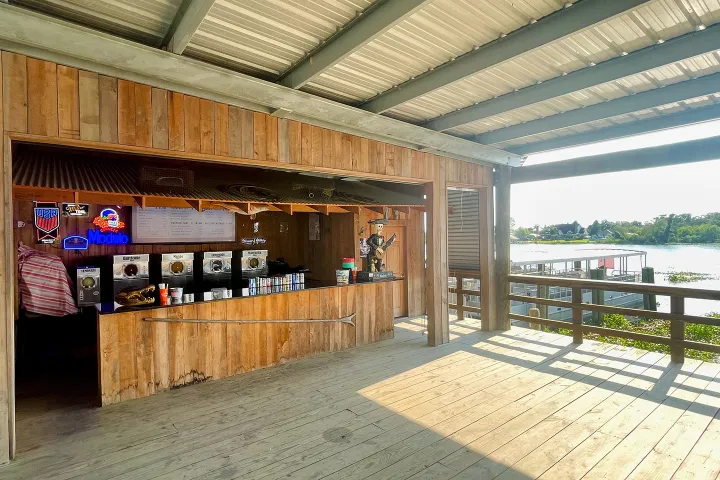 Wooden dockside bar with beverage machines and neon signs, overlooking water on a sunny day.