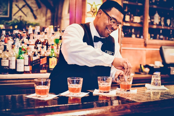 Bartender in uniform preparing cocktails at a bar with bottles in the background.
