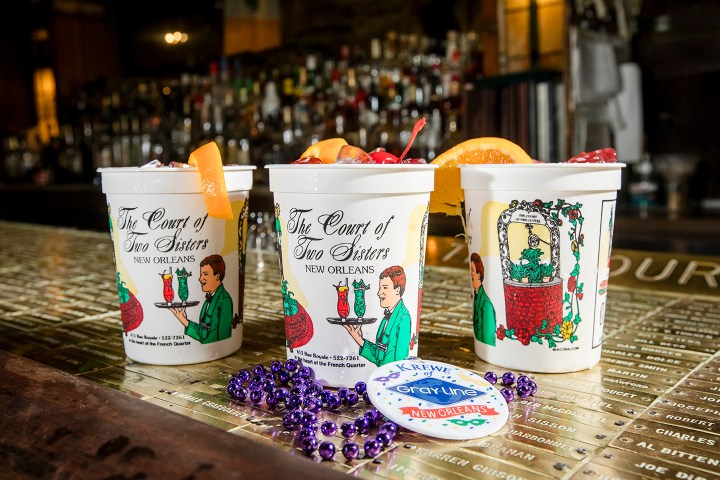 Three branded cups with drinks, garnished with orange slices, on a bar beside purple beads and a button.