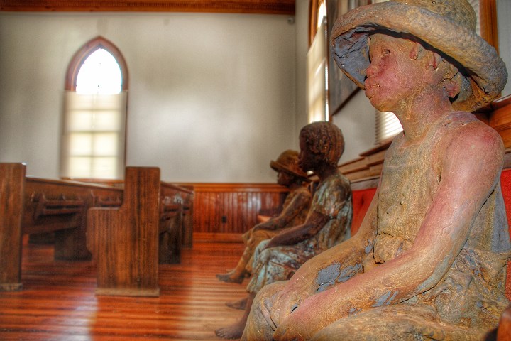 Clay statues of children in hats sitting inside a church with wooden pews and a stained glass window.