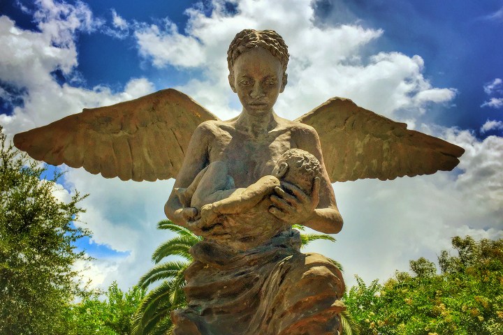 Angel statue with wings holding a baby against a cloudy sky background.