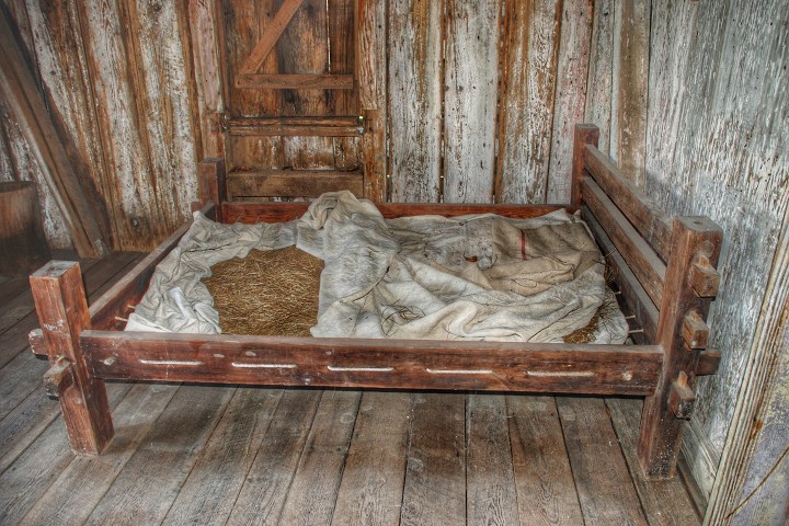 Old wooden bed with straw mattress and blankets in rustic room.