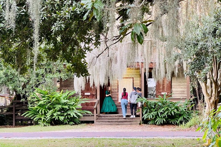 Four people on a porch of a rustic wooden house, surrounded by greenery and hanging moss.