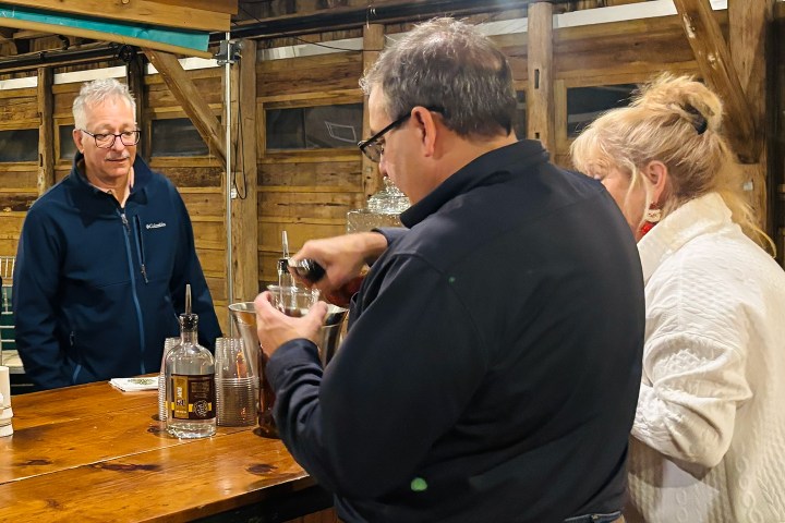 Two people pour drinks at a wooden bar while a man watches.