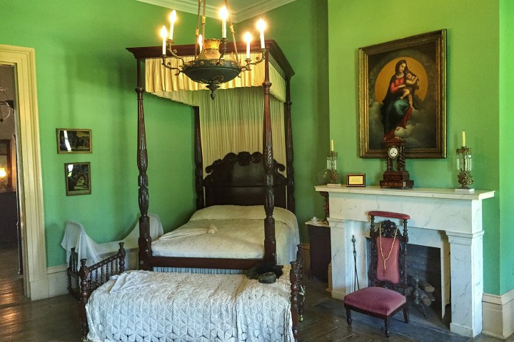 Vintage bedroom with canopy bed, green walls, chandelier, and a painting above a fireplace.