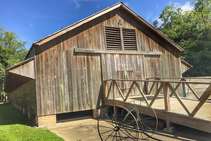 Wooden barn with a bridge and wheel in front, surrounded by trees and grass under a blue sky.