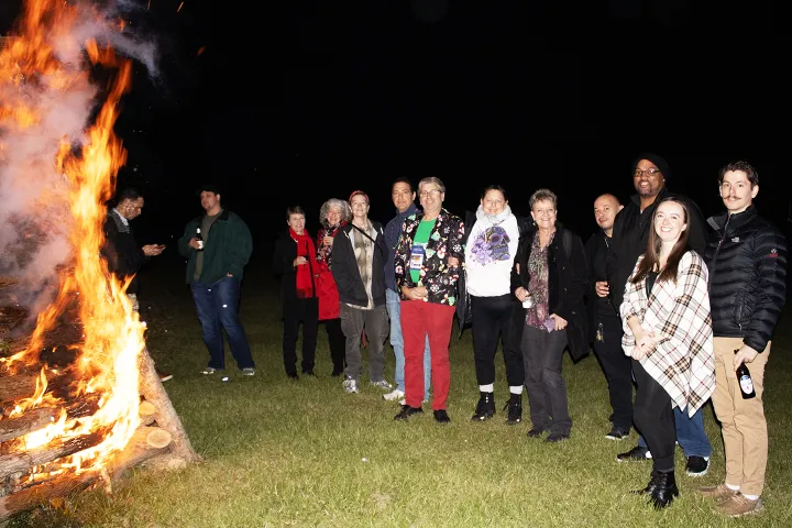Group of people standing near a large bonfire at night on a grassy area.