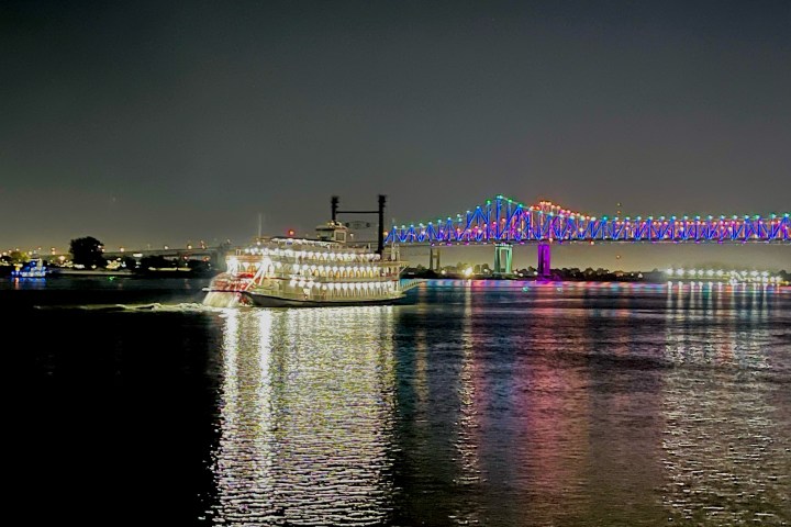 Lit-up riverboat on water at night near a colorful, illuminated bridge.