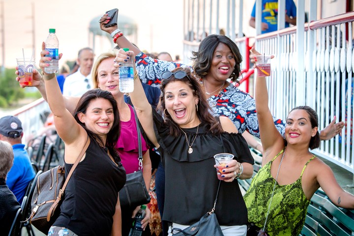 Five women joyfully holding drinks outdoors, raising their arms in celebration.